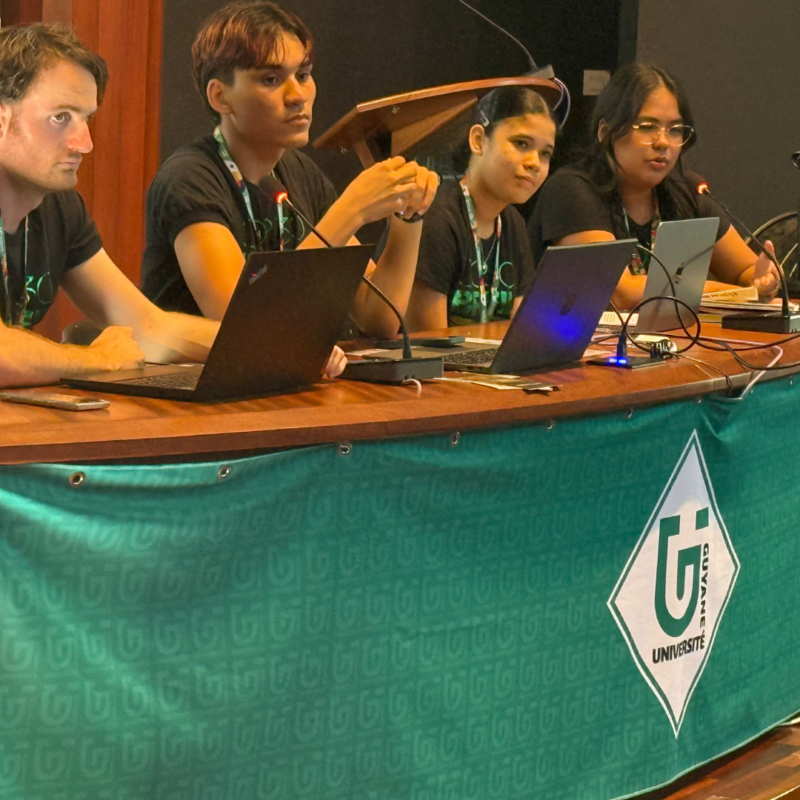 Quatre étudiants de l’Université de Guyane participent à une table ronde, assis derrière une table avec microphones et ordinateurs, lors d’un événement académique.
