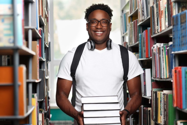 Étudiant souriant dans une bibliothèque, tenant une pile de livres.